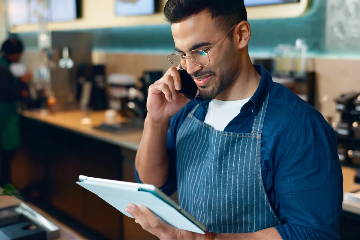 Homem, com avental de garçom ou chef, fala ao telefone enquanto checa informações em um tablet no balcão de um café, avaliando o impacto da escolha do regime tributário (Simples, Lucro Presumido ou Regime Específico) na Reforma Tributária.