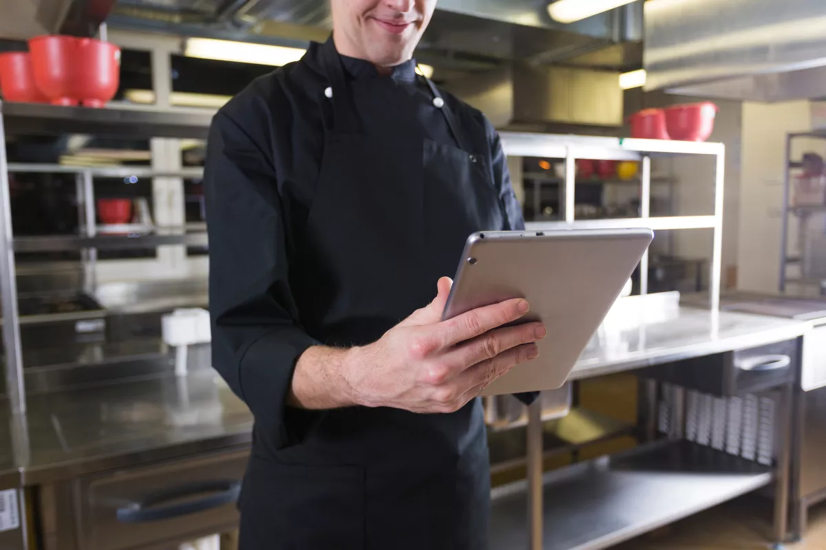 Chef em uniforme preto utilizando um tablet em uma cozinha industrial de aço inoxidável, inspecionando o cumprimento dos padrões de segurança alimentar, um dos indicadores de desempenho restaurantes.
