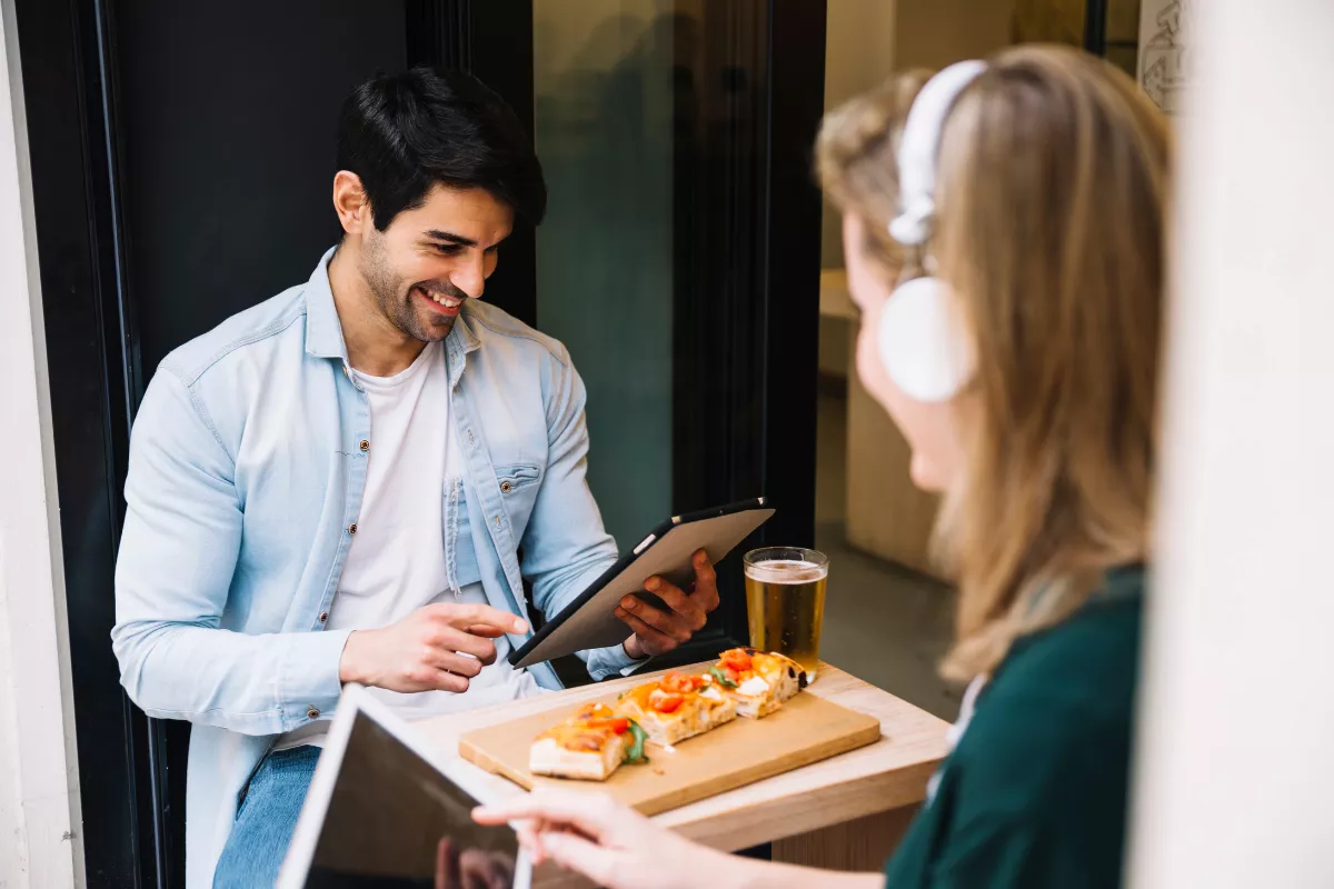 Cliente sorrindo e recebendo um prato de pizza em uma bandeja de um garçom que usa um tablet para registrar o tempo, ilustrando a eficiência na redução do Tempo de Ciclo do Pedido, um dos indicadores de desempenho restaurantes que impacta a satisfação.