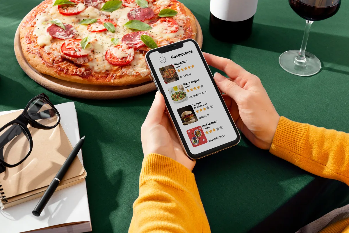 Pessoa fazendo pedido online em um cardápio digital pelo celular, ao lado de uma pizza, destacando como a presença digital e o delivery são fundamentais para aumentar as vendas no setor de alimentação