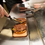 Profissional preparando cheeseburgers na Chapa Tradicional. A superfície lisa permite selar o pão e preparar diversos acompanhamentos ao mesmo tempo.