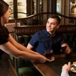Garçom sorridente entregando bebida a um cliente em uma mesa de estofado de couro, ilustrando atendimento gentil.