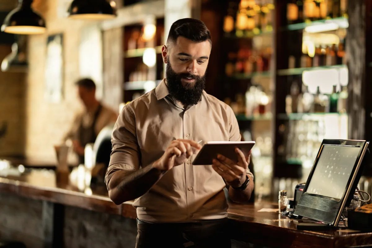 Barman utilizando tablet na frente de caixa, mostrando automação de pedidos e integração com o sistema PDV para bares e restaurantes