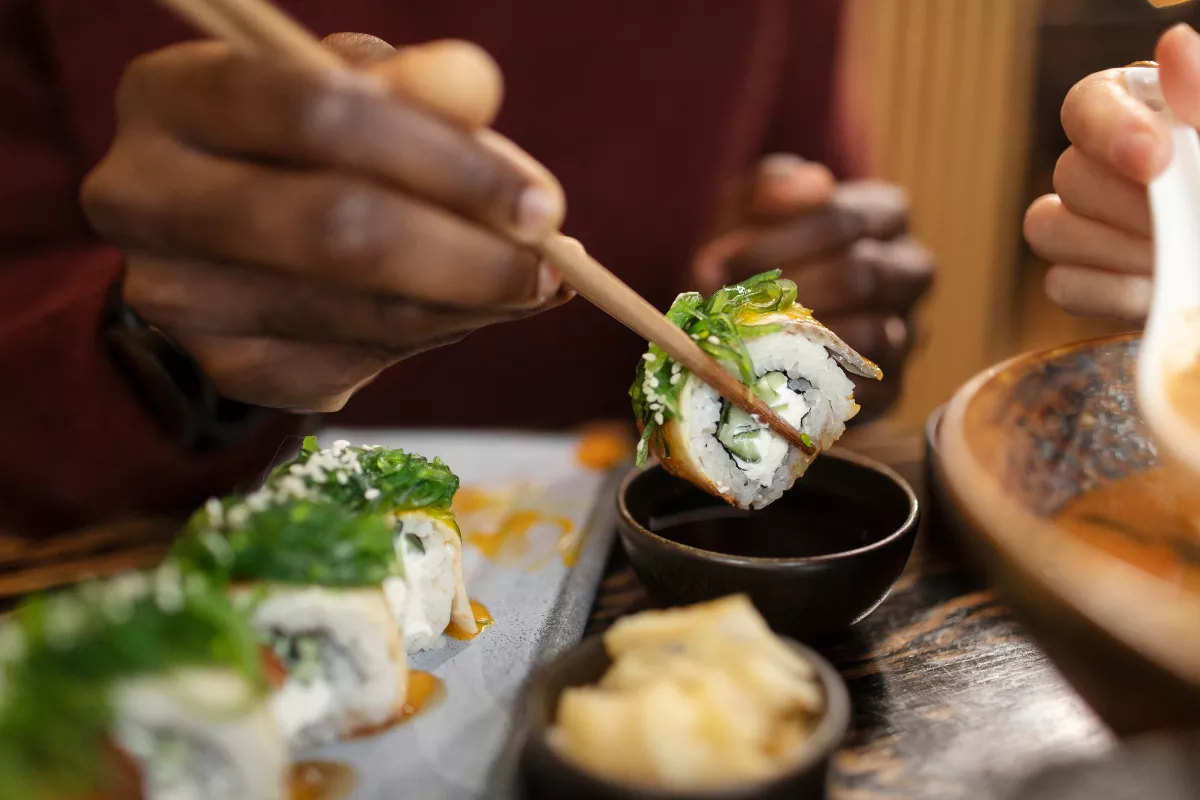 Close-up de uma pessoa utilizando hashi para pegar um uramaki de sushi (roll invertido) com alga marinha por cima. O controle do CMV e desperdício é crucial para a margem de lucro de restaurantes de culinária japonesa
