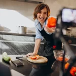 Proprietária de restaurante sorridente gravando vídeo e interagindo com o público, destacando a importância de produzir conteúdo digital para atrair clientes da região