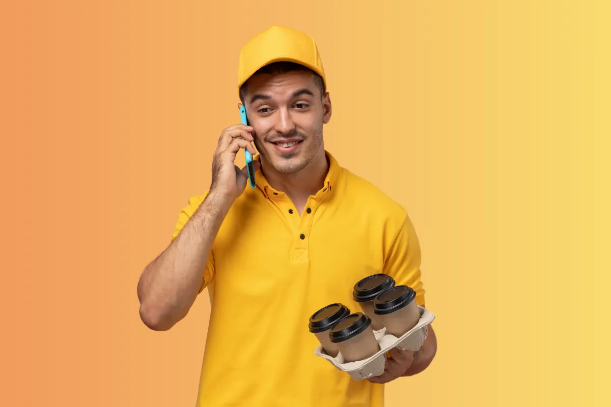 Jovem entregador de delivery, vestindo uniforme amarelo, sorrindo ao telefone e segurando quatro copos de café, representando a agilidade e os parceiros da 99Food.