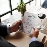 Visão superior de um homem em um terno lendo um cardápio de design elegante com o título 'Menu' e divisões como 'Starter', 'Main' e 'Dessert', ao lado de uma taça de vinho tinto e um copo d'água. A imagem destaca a apresentação de um menu degustação e a sofisticação da experiência, conectando-se à seção do texto que explica o conceito de 'tour gastronômico' e a desmistificação de que esse menu não é exclusivo de restaurantes de luxo