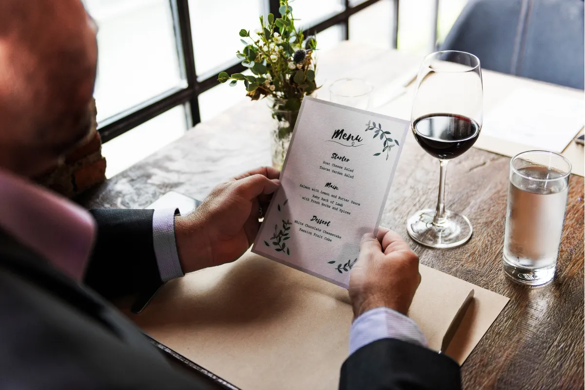 Visão superior de um homem em um terno lendo um cardápio de design elegante com o título 'Menu' e divisões como 'Starter', 'Main' e 'Dessert', ao lado de uma taça de vinho tinto e um copo d'água. A imagem destaca a apresentação de um menu degustação e a sofisticação da experiência, conectando-se à seção do texto que explica o conceito de 'tour gastronômico' e a desmistificação de que esse menu não é exclusivo de restaurantes de luxo