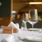 Mesa de restaurante elegante com toalha branca, copos de vinho e um cartaz de madeira escrito "RESERVED" (Reservado). Ao fundo, o salão do restaurante desfocado, indicando um ambiente preparado para clientes com reserva