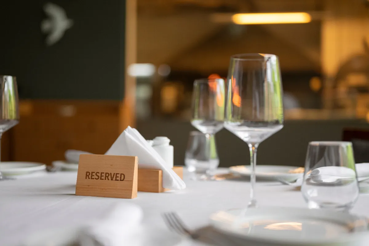 Mesa de restaurante elegante com toalha branca, copos de vinho e um cartaz de madeira escrito "RESERVED" (Reservado). Ao fundo, o salão do restaurante desfocado, indicando um ambiente preparado para clientes com reserva