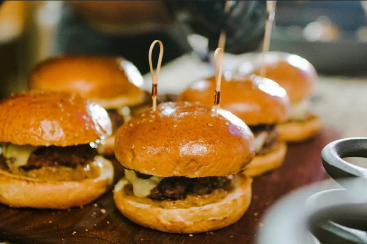 Close-up de uma sequência de mini-hambúrgueres apetitosos com queijo e molho, espetados com palitos decorativos, sobre uma tábua de madeira. Esta imagem ilustra perfeitamente a criatividade na aplicação do Menu Degustação em hamburguerias, conforme mencionado no texto, e a ideia de 'pequenas porções, grande experiência