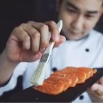 Sushiman com uniforme branco pincelando com molho o salmão fresco fatiado em cima de uma pedra escura. A qualidade da matéria-prima e do profissional afeta diretamente o lucro do restaurante japonês