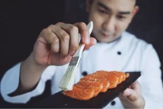 Sushiman com uniforme branco pincelando com molho o salmão fresco fatiado em cima de uma pedra escura. A qualidade da matéria-prima e do profissional afeta diretamente o lucro do restaurante japonês