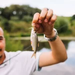 Homem segurando um pequeno peixe recém-pescado, à beira de um lago em um pesque e pague, ilustrando a experiência do cliente nesse tipo de estabelecimento e a importância de registrar e pesar cada pescado de forma eficiente.