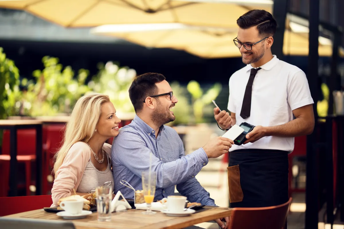 Garçom sorridente em um restaurante ao ar livre, segurando uma maquininha de cartão e um smartphone. Um casal feliz está pagando a conta em uma mesa, evidenciando um atendimento eficiente e moderno, possivelmente otimizado por um sistema como o CNM