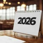 Calendário de mesa com o ano 2026 em destaque sobre uma superfície de madeira e fundo desfocado de restaurante, simbolizando o planejamento de marketing e datas comemorativas.