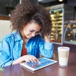 ovem sorridente em cafeteria utilizando um tablet sobre a mesa para navegar pelo cardápio digital, ilustrando a autonomia e facilidade que a tecnologia proporciona na escolha do pedido em restaurantes e cafés.