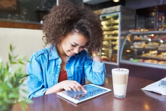 ovem sorridente em cafeteria utilizando um tablet sobre a mesa para navegar pelo cardápio digital, ilustrando a autonomia e facilidade que a tecnologia proporciona na escolha do pedido em restaurantes e cafés.