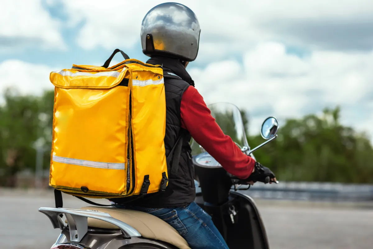 Entregador de delivery visto de costas em uma moto com uma mochila térmica amarela, representando a logística de entregas e a necessidade de integração de múltiplos canais de venda em um único sistema.
