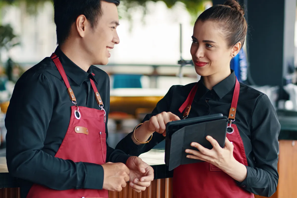 Dois garçons de avental vermelho conversando e olhando para um tablet, gerenciando pedidos digitais de forma integrada para garantir agilidade e comunicação entre salão e cozinha.