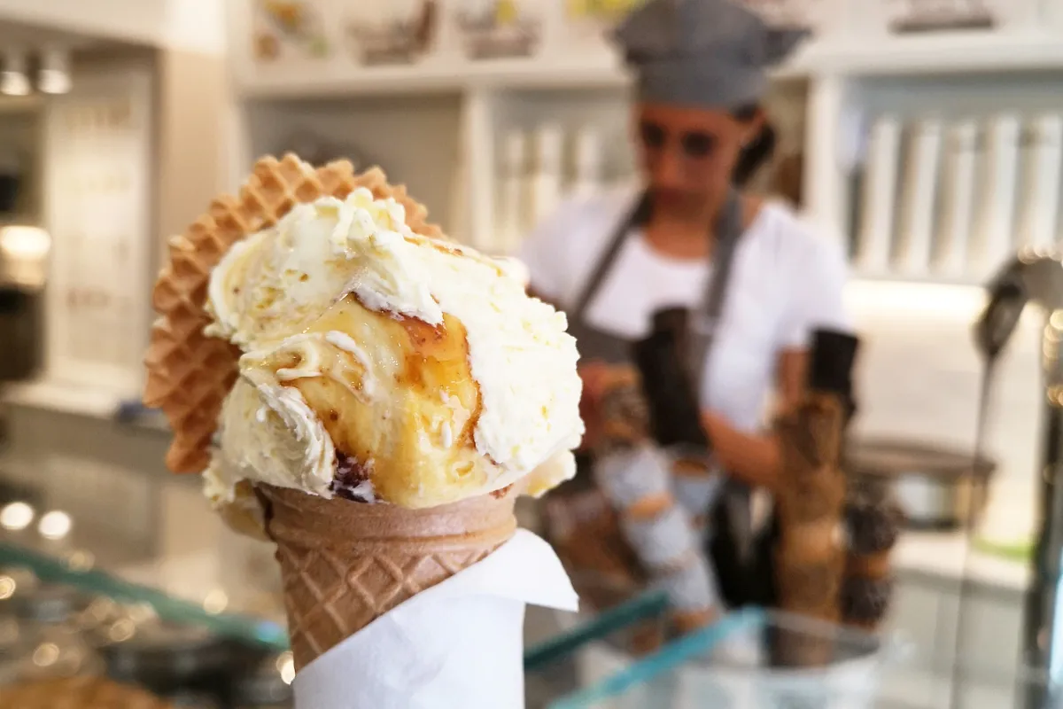 Close-up de uma casquinha de sorvete artesanal com calda, o principal produto de venda, com uma funcionária uniformizada trabalhando ao fundo da operação da sorveteria.