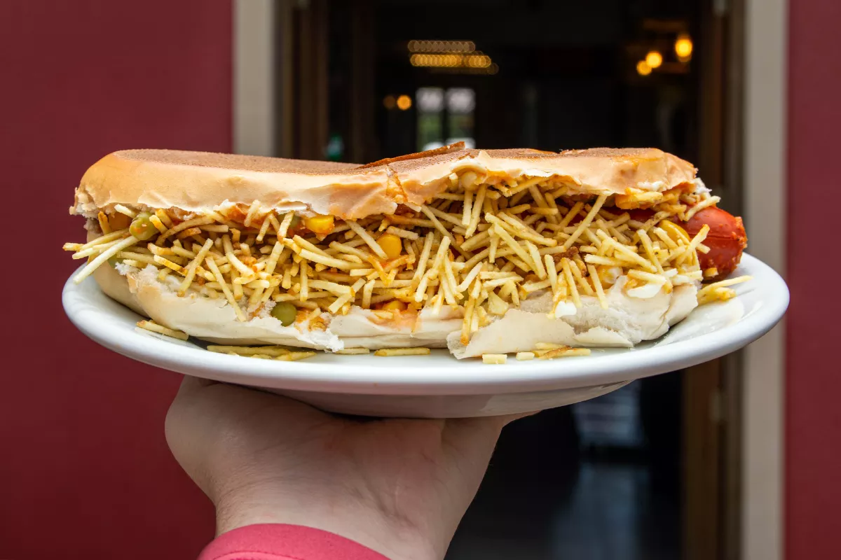 Cachorro-quente recheado com batata palha servido em prato branco, ilustrando o produto típico de carrinhos de cachorro-quente e o potencial de lucro no setor de alimentação de rua