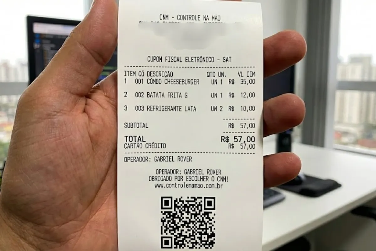 Cupom fiscal eletrônico SAT emitido pelo sistema CNM, sendo segurado na mão de uma pessoa. O cupom apresenta detalhes de uma venda em restaurante, com discriminação dos itens vendidos, valores, forma de pagamento, operador de caixa e QR Code para validação. Exemplo de documento fiscal simplificado que comprova a compra realizada pelo cliente no food service.