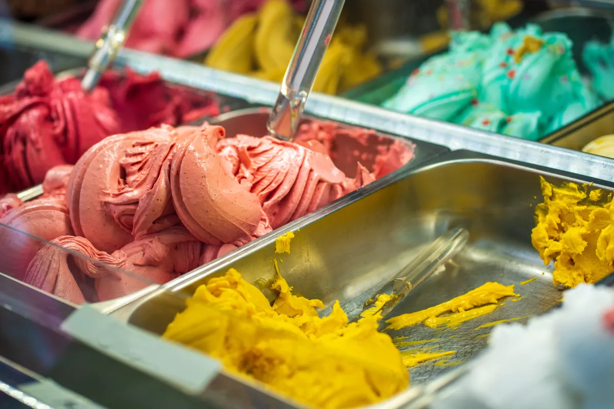 Vitrine de uma sorveteria pequena repleta de cubas com sabores variados e vibrantes de sorvete (vermelho, rosa, amarelo e azul), com pás de servir prontas para o atendimento ao cliente.