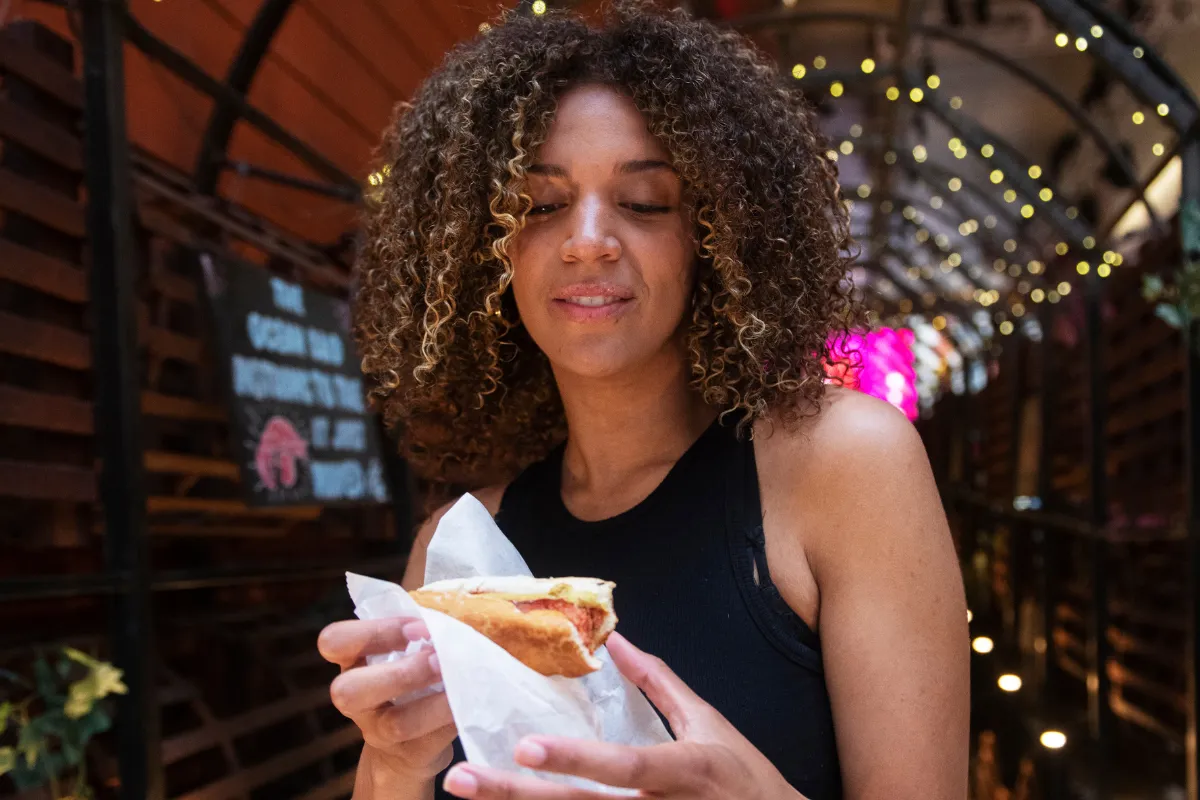 Mulher jovem e sorridente segurando um cachorro-quente gourmet em um ambiente com luzes decorativas ao fundo. A satisfação do cliente com a qualidade do produto é um dos principais fatores que influenciam o resultado financeiro de uma lanchonete.