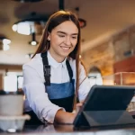 Atendente de cafeteria sorrindo ao usar um tablet como frente de caixa intuitivo para registrar pedidos, ilustrando a facilidade de uso e modernização do atendimento com o sistema CNM.