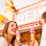 Duas jovens clientes sorrindo enquanto consomem casquinhas de sorvete rosa em ambiente externo, representando o público-alvo e o movimento que gera o faturamento mensal.