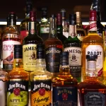 Prateleira de bar abastecida com uma ampla variedade de garrafas de whisky e bourbon, como Jameson e Jim Beam, ilustrando um estoque robusto e o investimento em mercadorias para uma distribuidora de bebidas de maior porte.