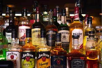 Prateleira de bar abastecida com uma ampla variedade de garrafas de whisky e bourbon, como Jameson e Jim Beam, ilustrando um estoque robusto e o investimento em mercadorias para uma distribuidora de bebidas de maior porte.
