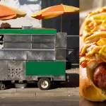 Carrinho de cachorro-quente de rua com guarda-sóis e detalhe de hot dog com molho e mostarda, ilustrando o ponto de venda e os fatores que influenciam o lucro desse tipo de negócio