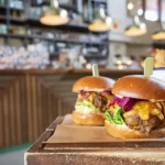 Dois hambúrgueres artesanais gourmet bem montados sobre uma mesa de madeira, com o balcão da lanchonete ao fundo. A venda de produtos de alto valor agregado é fundamental para aumentar o ticket médio e definir quanto lucra uma lanchonete delivery.