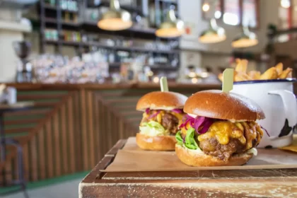 Dois hambúrgueres artesanais gourmet bem montados sobre uma mesa de madeira, com o balcão da lanchonete ao fundo. A venda de produtos de alto valor agregado é fundamental para aumentar o ticket médio e definir quanto lucra uma lanchonete delivery.