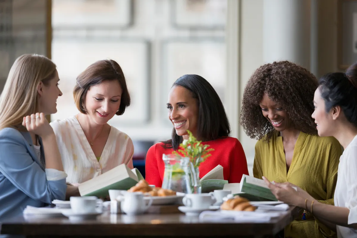 Grupo de mulheres sentadas à mesa de um café segurando livros e conversando animadamente, representando ações culturais como o Dia Nacional da Leitura em outubro.