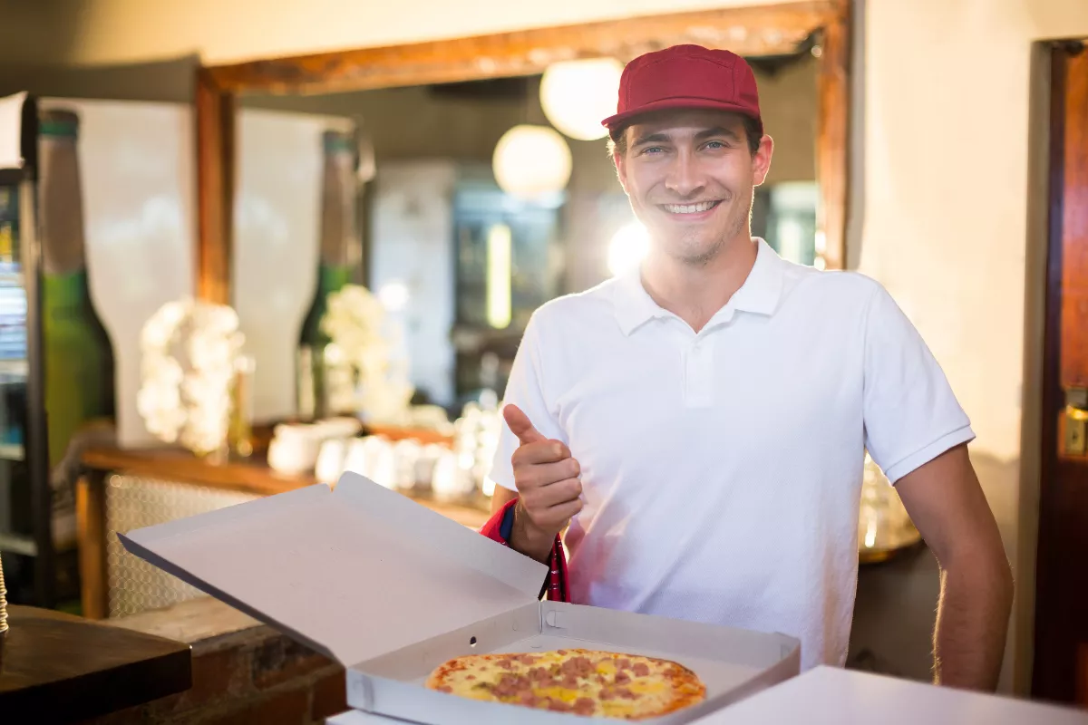 Entregador de pizza sorridente de boné vermelho fazendo sinal de positivo ao lado de caixas de pizza abertas, representando a agilidade no delivery e a boa experiência do cliente como parte fundamental da propaganda