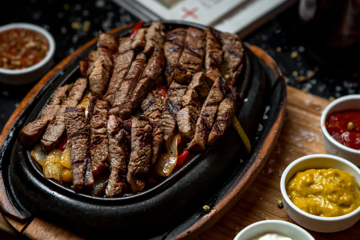 Porção de carne fatiada, pronta para ser servida em uma chapa quente com molhos. A imagem foca na apresentação do prato final ao cliente e na padronização das porções