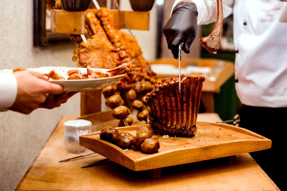 Chef profissional servindo uma costela assada com batatas em uma tábua de madeira, ilustrando um prato de churrasco bem apresentado em um restaurante. A imagem remete ao controle de porções e custos.