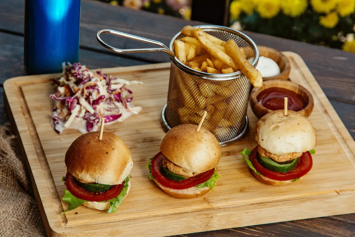 Tábua de madeira servida com três mini burgers, cesta de batata frita e porção de salada coleslaw, uma ótima opção de petiscos para um Combo Happy Hour.