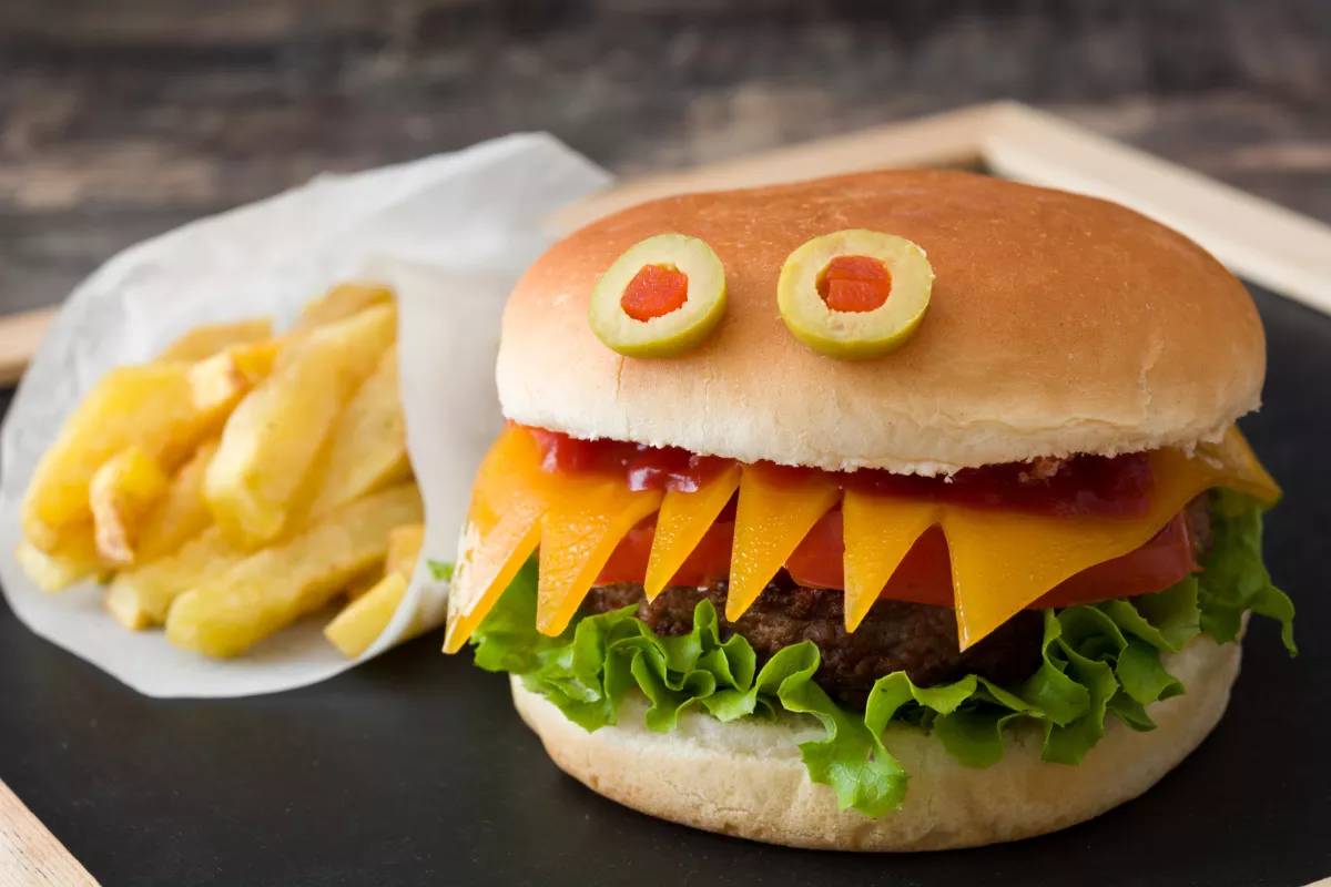 Hambúrguer divertido decorado como um monstrinho com dentes de queijo e olhos de azeitona, ao lado de batatas fritas, focado em atrair crianças no Combo Kids.