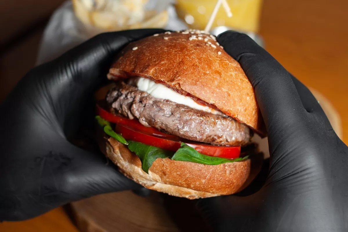 Mãos com luvas pretas segurando com destaque um hambúrguer gourmet, representando a curiosidade e exclusividade de um Combo Misterioso ou Secreto.