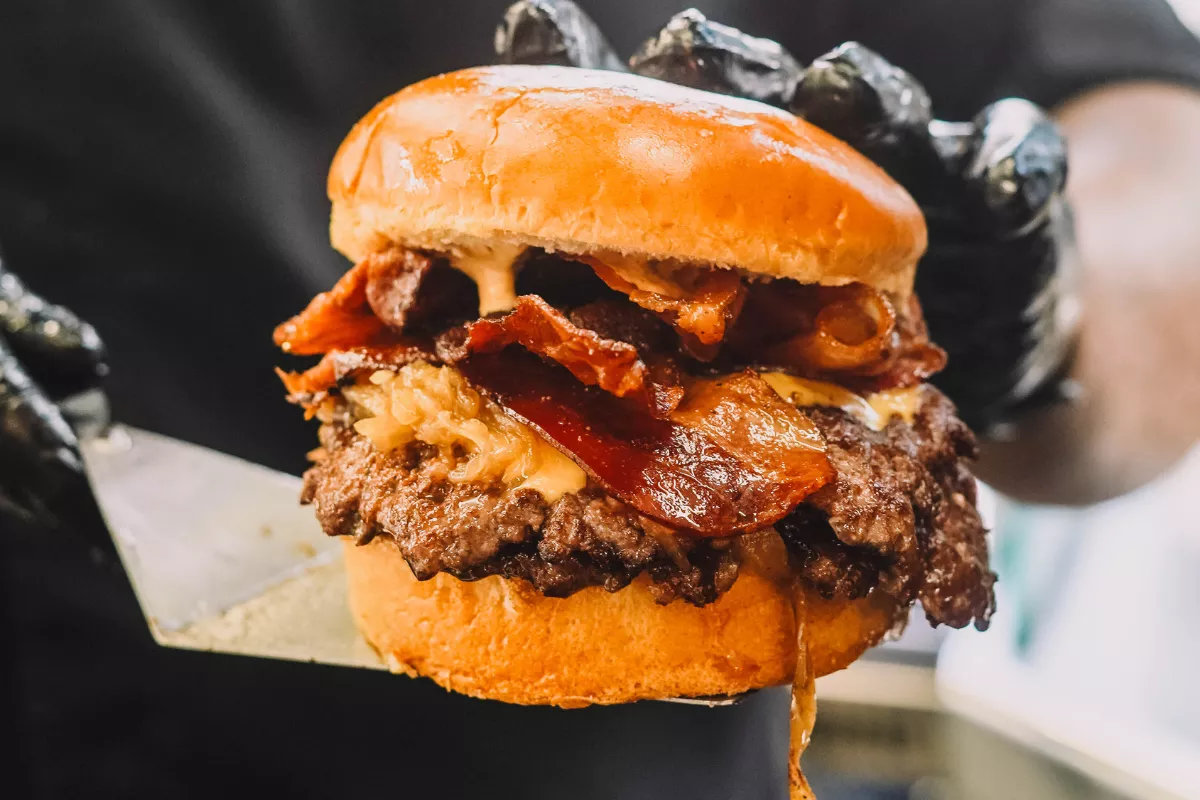 Mãos com luvas pretas segurando uma espátula com um hambúrguer grande e suculento, recheado com bastante bacon e cebola caramelizada, ilustrando a opção de Combo Larica ou Matador de Fome.
