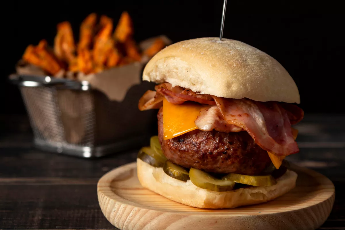 Hambúrguer artesanal montado com bacon e picles em prato de madeira, acompanhado de cesta de batata doce frita ao fundo, exemplificando a liberdade de escolha do Combo Monte o Seu.