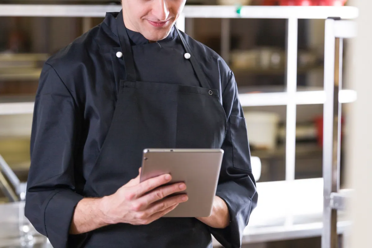 Chef de cozinha com uniforme preto utilizando um tablet para visualizar pedidos, demonstrando a automação da operação do restaurante através de sistemas como KDS e comanda eletrônica.