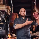 Chef segurando um corte de carne crua, tipo T-bone, em uma cozinha comercial. A imagem destaca a importância da seleção de insumos e a pesagem da carne crua para a ficha técnica