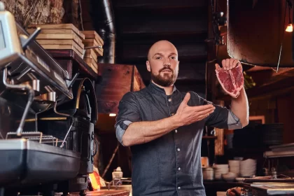 Chef segurando um corte de carne crua, tipo T-bone, em uma cozinha comercial. A imagem destaca a importância da seleção de insumos e a pesagem da carne crua para a ficha técnica