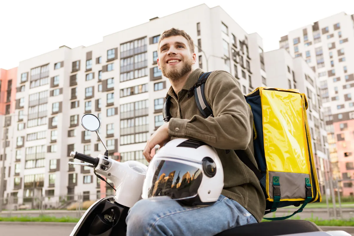 Um jovem entregador sorridente sentado em sua moto branca, segurando um capacete branco no colo. Ele usa uma mochila de entrega amarela e está posicionado em frente a prédios residenciais, transmitindo profissionalismo e a confiança necessária ao buscar serviços terceirizados de entrega.