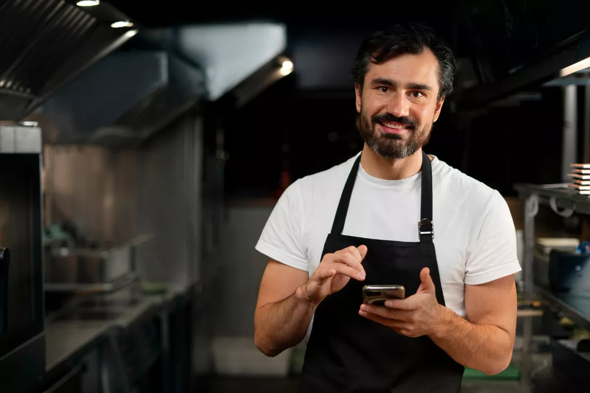 Dono de hamburgueria sorrindo e usando um smartphone dentro da cozinha profissional, ilustrando a facilidade de gerenciar combos, estoque e operação pelo celular com um sistema de gestão eficiente.